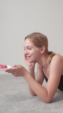 Dikey video. Fiziksel aktivite. Lezzetli bir mola. Siyah spor giysili güzel, gülümseyen bir bayan. Sabahları evde elinde pastayla tabağa bakıyor..