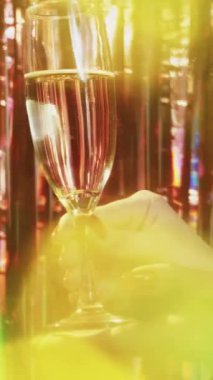 Dikey video. Şampanya partisi. Şenlikli bir kutlama. Işıl ışıl ışıl arka planda renkli mercekli bir bardak köpüklü alkollü kadın el feneri..