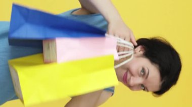 Dikey video. Heyecan verici alışveriş. Perakende terapisi. Gülümseyen kadın, sarı boş alanda izole edilmiş renkli kağıt torbalarla gösteriş yapmaktan mutlu..