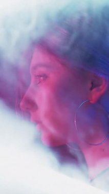 Dikey video. Neon ışıklı surat. Siber punk güzeli. Disko 80 'lerin gece hayatı. Pembe mavi renkli parlak profil. Koyu renkli beyaz duman bulutu içinde bir kız..