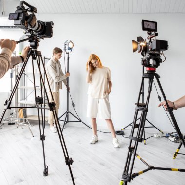 Model hazırlığı. Video kamera stüdyosu. Sanat ekibi fotoğraf için ekipmanları ayarlıyor. Kız, operatörlerin kameraların ışıklandırma seviyesini ayarlamasına yardım ediyor.