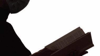 Karantina çalışması. Siluet okunuyor. Salgın hobisi. Yüz maskesi takmış, açık kitabı beyaz boş arkaplanda izole bir kadının koyu renk kontrast profili..