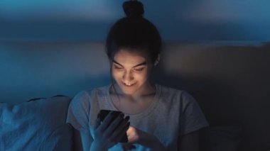 Gece online. Uyku zamanı aleti. Neşeli, rahat bir kız telefonunu kullanıyor internette eğleniyor. Gece geç saatlerde yatakta internette geziniyor. Karanlık yatak odasında..