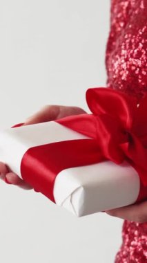 Dikey video. Xmas hediyesi. Tatil sürprizi. Parlak elbiseli, kırmızı kurdeleyle sarılı hediye kutusunu tutan, beyaz elbiseli, tanınmayan zarif bir kadın..