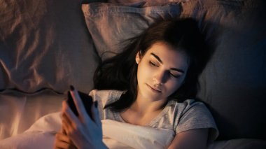 Gece sohbeti. Geç iletişim. Çevrimiçi boş zaman. Rahat, meraklı bir kadın. Karanlık yatak odasında cep telefonuyla mesaj atıyor..