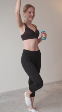 Dikey video. Spor ağırlığı. Zayıflık kadını. Gülümseyen süs kızı evde dambılları kaldırıyor. Gücünü ve forma bağlılığını gösteriyor..