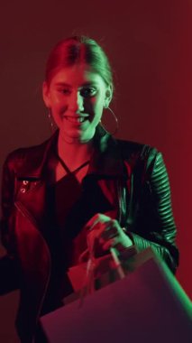 Dikey video. İyi alışverişler. Kara Cuma. İnternetten alışveriş. Gece satışı. Kırmızı yeşil renkli neon ışıklı kız, satın alma çantalarıyla, koyu renk 2 'li sette gülümsüyor..