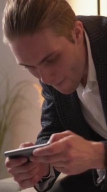 Dikey video. İnternet bağımlılığı. Dijital teknoloji. Modern takım elbiseli, elinde akıllı telefon olan yakışıklı bir adam internette aletin üzerinde oturuyor..