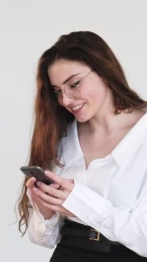 Dikey video. Mobil sohbet. Çevrimiçi iletişim. Gözlüklü mutlu bir kadın, beyaza izole edilmiş telefon kullanarak mesaj okumaktan zevk alıyor..