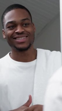 Dikey video. Erkek bakımı. Yüz bakımı. Beyaz havluyla tatmin olmuş, aynaya bakan, sakalını tıraş ettikten sonra yüzüne dokunan adam..