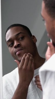 Dikey video. Erkek derisi. Sivilce tedavisi. Deri rutini. Beyaz havluyla kendine güveni olan adam aynaya baktıktan sonra yüzüne dokunuyor..