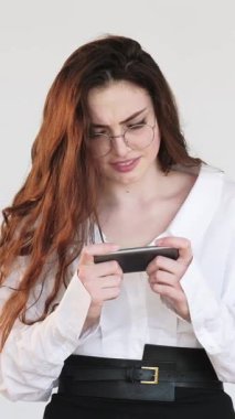 Dikey video. Mobil oyun. Gadget eğlencesi. Heyecanlı, gözlüklü akıllı kadın beyazlar içinde izole bir şekilde internette oynamaktan zevk alıyor..