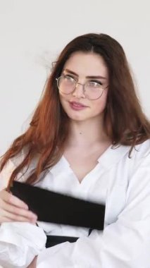 Dikey video. Ofis güzeli. Flört eden yüz. Utangaç, gözlüklü, el panosu sallayan iş kadını. Serinlemek için yelpaze gibi..