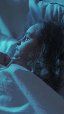 Dikey video. Çocuk korkusu. Gece terörü. Uyku sorunu. Korkmuş küçük bir kız battaniyenin altında saklanıyor. Tek başına, karanlık bir odada, mavi ay ışığında yatıyor..