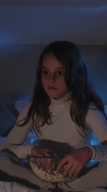 Dikey video. Yatma vakti. Çocuk eğlencesi. Korkmuş, korkmuş, şok olmuş, patlamış mısır yemekten zevk alan bir kız. Gecenin karanlığında, tek başına, mavi ışıklı bir evde oturuyor..
