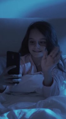 Dikey video. Gece çevrimiçi arama. Çocuk sohbeti. Geç iletişim. Neşeli küçük kız cep telefonu kullanıyor. El sallıyor. Evdeki karanlık odada mavi ışıkla oturuyor..