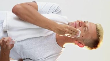 Dikey video. Yüz temizleme. Cilt bakımı. Sivilce tedavisi. Kendine güvenen sarışın adam beyaz pamuk pedi kullanarak yüzünü tonikle temizliyor..