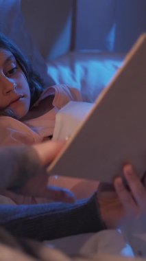 Dikey video. Gece hikâyesi. Yatmadan önce kitap oku. Çocuk uykusuzluğu. Ebeveyn yaşam tarzı. Anne kitaptaki resimlerini, yatak odasında yatan yorgun küçük kıza gösteriyor..