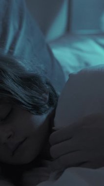 Dikey video. Uyuyan çocuk. Anne bakımı. Çocuk yatma vakti. Kadın elleri battaniyeyle örtülmüş. Huzurlu küçük kızı okşuyor. Geceleri yatak odasında mavi ay ışığıyla yatıyor..