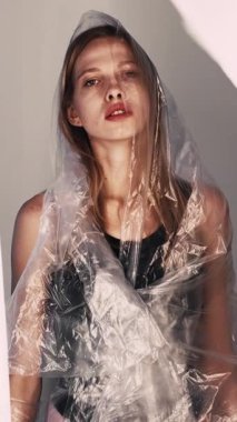 Dikey video. Zihinsel bozukluk. Kadın depresyonu. Gri ışıklı sanatta acı çeken şeffaf polietilen film giyen yorgun kadın..