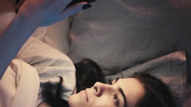 Dikey video. Gadget gecesi. İnternette uykusuzluk. İnternet bağımlılığı. Uykucu sıkılmış kız, karanlık yatak odasında cep telefonunu kullanıyor..