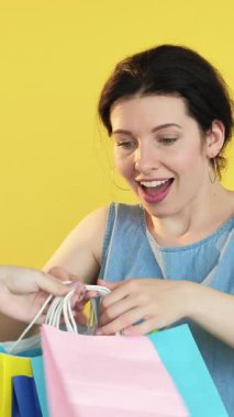 Dikey video. Alışveriş kavgası. Kara Cuma. Satın almayı reddeden mutlu kadın satın alma yarışması sarı arkaplanda izole edilmiş başka bir müşteriye renkli kağıt torbaları veriyor..