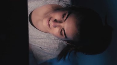 Dikey video. Geç saatlere kadar sohbet. Gece online. İnternet iletişimi. Mutlu neşeli kız karanlık yatak odasında dizüstü bilgisayar kullanarak mesaj yazıyor..