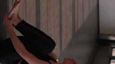 Dikey video. Jimnastik pratiği. Pilates egzersizi. Spor kıyafetli sağlıklı bir kadın spor salonunda spor salonundaki yoga minderinde ayak tutarak bacak kaldırma egzersizi yapıyor..