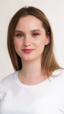 Dikey video. Mutlu genç portre. Kadınsı cazibe. Makyajlı, beyaz tişörtlü, saçı dökülmüş, arka planda açık bir şekilde duran iyimser utangaç kız..