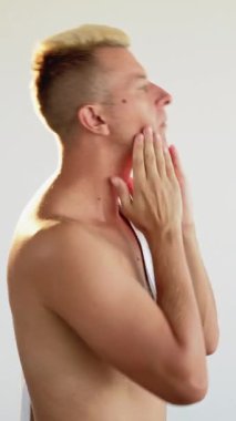 Dikey video. Cilt bakımı. Yüz bakımı. Tuvalet rutini. Tıraş olduktan sonra nemlendirici yüz kremi süren kendinden emin, sarışın bir adamın profili..