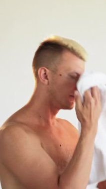 Dikey video. Sabah hijyeni. Tımar rutini. Tatmin olmuş, üstsüz, sarışın bir adam. Beyaz bir banyodan sonra beyaz havluyla yüzünü siliyor..