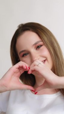 Dikey video. Aşk sinyali. Romantik mesaj. Takdir, sevgiyi destekler. Beyaz tişörtlü neşeli genç kız ışık fotokopi arka planında izole edilmiş kalp hareketi gösteriyor..