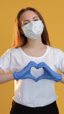 Dikey video. Aşk sinyali. Karantina ilişkisi. Korunaklı yüz maskeli kadın. Eldivenler, sarı renkte kalp hareketi gösteriyor..