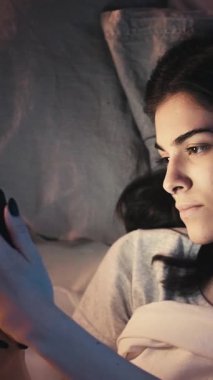 Dikey video. Gadget gecesi. İnternette uykusuzluk. İnternet bağımlılığı. Uykucu sıkılmış kız, karanlık yatak odasında cep telefonunu kullanıyor..