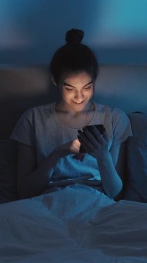 Dikey video. Gece online. Uyku zamanı aleti. Neşeli, rahat bir kız telefonunu kullanıyor internette eğleniyor. Gece geç saatlerde yatakta internette geziniyor. Karanlık yatak odasında..