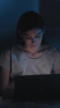 Dikey video. Gece işi. Serbest bir iş. Uykusuzluk teknolojisi. Gece geç saatlerde yatakta bilgisayarla yazı yazan rahat bir kadın. Karanlık ev yatak odası..
