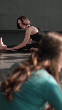Dikey video. Esneme stüdyosu. Pilates egzersizi. Grup üyeleri spor salonundaki spor minderi üzerindeki kadın yoga eğitmeninden sonra jimnastik sınıfı egzersizlerini tekrarlıyor..