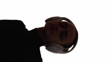 Dikey video. Dans eden siluet. Müzik zevki. Kulaklıklı heyecanlı kadının, beyaz fotokopi arkaplanından izole edilmiş ses aygıtını dinlemenin keyfini çıkardığı koyu renk kontrast çizgisi..