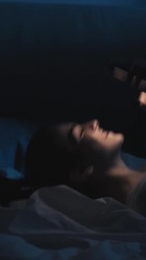Dikey video. Romantik sohbet. Gece online. Randevu uygulaması. Rahatlamış mutlu aşık bir kadın telefonda gülümseyen bir mesaj okuyor geç saatte yatakta, karanlık odada..