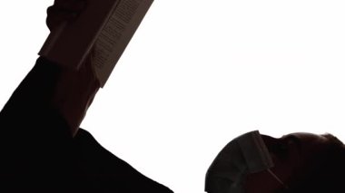 Dikey video. Karantina çalışması. Siluet okunuyor. Salgın hobisi. Yüz maskesi takmış, açık kitabı beyaz boş arkaplanda izole bir kadının koyu renk kontrast profili..