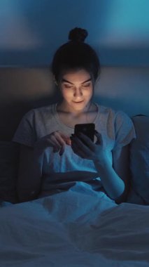 Dikey video. Gadget gecesi. Ev interneti. İnternette uykusuzluk. Karanlık yatak odasında geç saatte cep telefonunu kullanan meraklı kız..