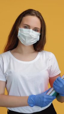 Dikey video. Karantina eğitimi. Sosyal uzaklık. COVID-19 salgın enfeksiyonu önleme koruyucu maske eldivenli kadın öğretmeni rahatlattı ve sarı üzerine izole kitaplar bıraktı..