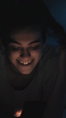 Dikey video. Gece sohbeti. Geç iletişim. Çevrimiçi boş zaman. Mutlu şakacı kadın mesajlaşır mesajlaşır. Karanlık odada cep telefonuyla flört eder..