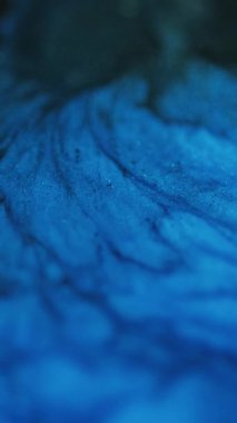 Dikey video. Islak doku. Parıltılı boya akışı. Karanlık soyut sanat arka planında odaksız mavi renkli parıldayan parçacıklar akrilik mürekkep dalgası hareketi.