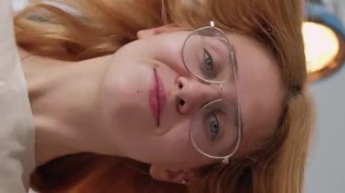 Dikey video. Fotoğraf çekimi. Kadın pozu. Gülümseyen sarışın kız kameraya göz kırpıyor. Stüdyoda gözlüklü neşeli, kendine güvenen bir güzellik..