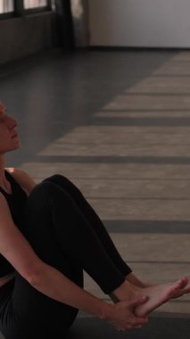 Dikey video. Jimnastik pratiği. Pilates egzersizi. Spor kıyafetli sağlıklı bir kadın spor salonunda spor salonundaki yoga minderinde ayak tutarak bacak kaldırma egzersizi yapıyor..