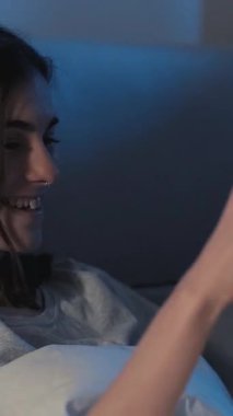 Dikey video. Gece okumaları. Yatak hobisi. Neşeli, meraklı kız kitap eğlenceli hikayesiyle eğleniyor. Gece geç saatlerde ev odasının karanlığında gülüyor..