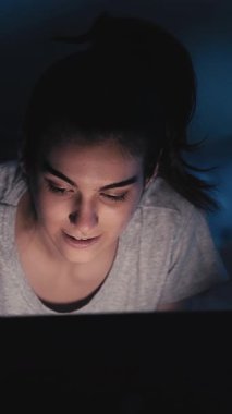 Dikey video. Geç saatlere kadar sohbet. Gece online. İnternet iletişimi. Mutlu neşeli kız karanlık yatak odasında dizüstü bilgisayar kullanarak mesaj yazıyor..