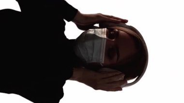 Dikey video. Karantina eğlencesi. Salgın müzik. Yüzü maskeli, koyu renkli bir kadın silueti. Beyazların izole edilmiş kulaklıklarındaki sesi dinlemekten zevk alıyor..