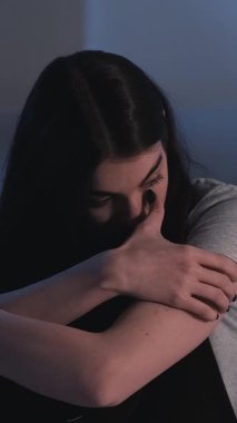 Dikey video. Gece depresyonu. Uykusuzluk yorgunluğu. Yorgun, rahatsız, endişeli, yalnız bir kadın karanlık bir yatak odasında dizlerine sarılıyor..
